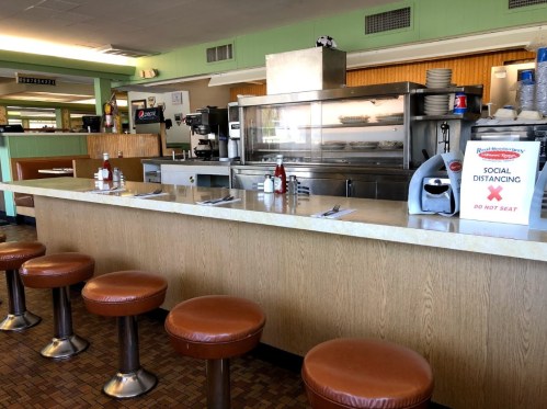 A diner interior with empty stools at the counter, a sign for social distancing, and a display of food behind glass.