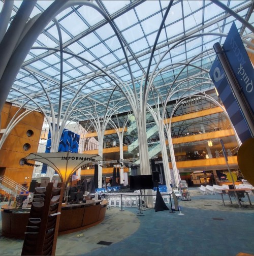 Modern atrium with a glass roof, featuring white structural beams and an information desk in a spacious, open area.