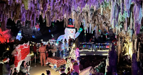 A festive cave scene with colorful lights, a large snowman, and visitors in holiday attire enjoying the decorations.