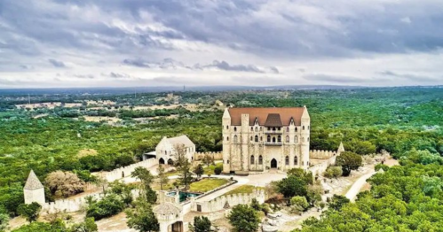 Aerial view of a large stone castle surrounded by lush greenery and trees under a cloudy sky.