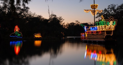 Colorful holiday lights depict a ship and a mermaid, reflecting on a calm water surface at dusk.