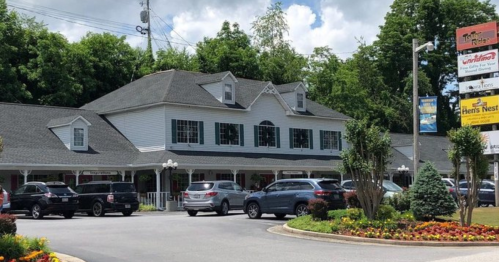 A white two-story building with a gray roof, surrounded by trees and parked cars, with a signpost nearby.