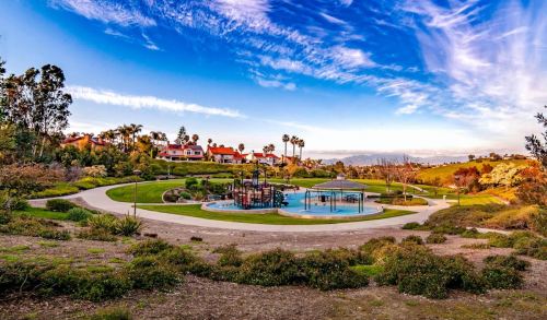 A scenic park with a playground, walking paths, and lush greenery under a blue sky with wispy clouds.