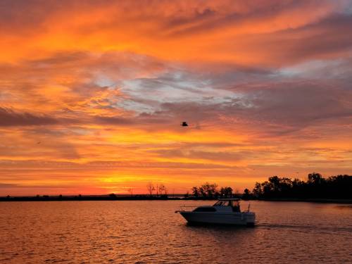 A boat glides across a calm waterway at sunset, with vibrant orange and purple clouds reflecting in the sky.