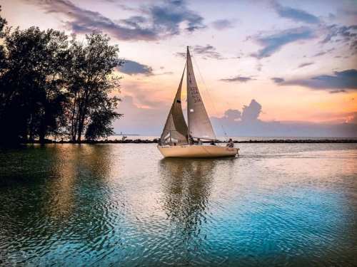 A sailboat glides across calm waters at sunset, surrounded by trees and a colorful sky.