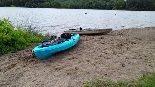 Two kayaks on a sandy beach by a lake, with people swimming in the water and greenery in the background.