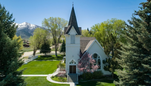 A quaint white church with a steeple, surrounded by green trees and a mountain backdrop under a clear blue sky.