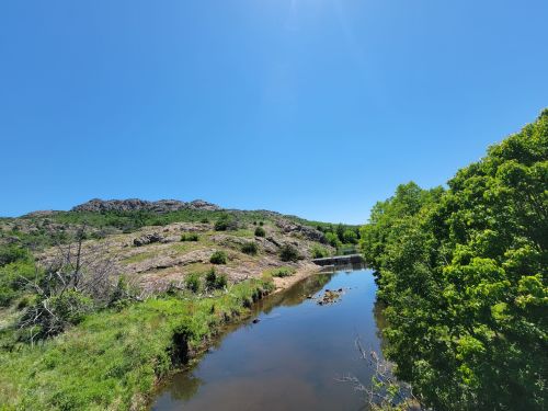 A serene landscape featuring a calm river, lush greenery, and rocky hills under a clear blue sky.