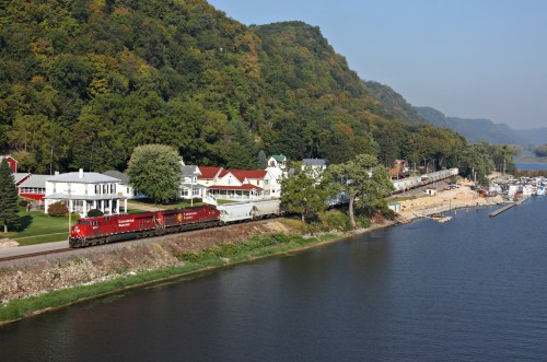 A train travels along a riverbank, with lush hills and a quaint town in the background.