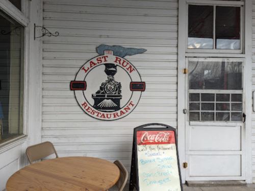Sign for "The Last Run Restaurant" featuring a train logo, with a menu board outside and a table nearby.