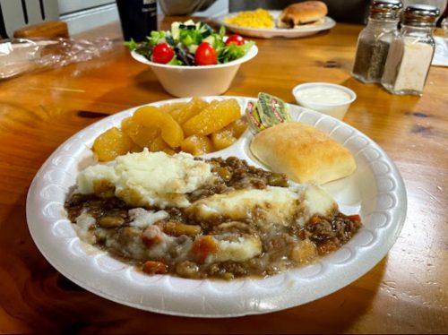 A plate with mashed potatoes, beef stew, peach slices, a roll, and a side salad with cherry tomatoes.