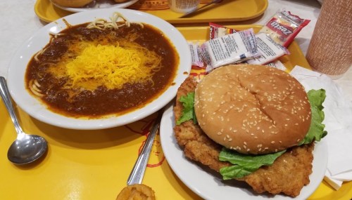 A plate with a fried chicken sandwich and a bowl of chili topped with shredded cheese, served on a yellow tray.