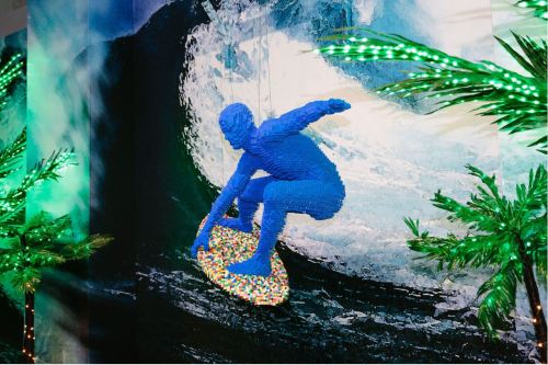 A blue LEGO figure surfing on a colorful board in front of a wave, surrounded by palm trees and vibrant lights.