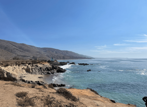 A serene coastal view with rocky shores, gentle waves, and distant hills under a clear blue sky.
