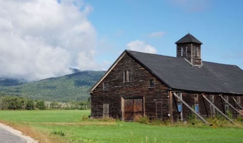 A rustic wooden barn stands beside a green field, with mountains and cloudy skies in the background.