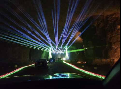 Colorful laser lights illuminate a dark cave, with cars parked along a winding path.