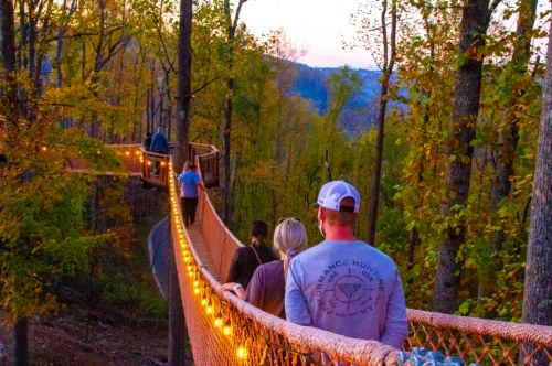 A group of people walking on a lit suspension bridge through a colorful autumn forest at sunset.