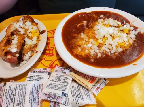 A plate with a chili dog topped with cheese and onions, next to a bowl of chili with cheese and onions, surrounded by condiment packets.