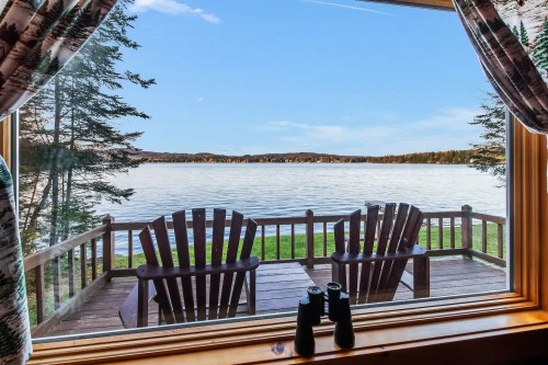 View from a cabin window overlooking a serene lake, with two Adirondack chairs and binoculars on a wooden deck.