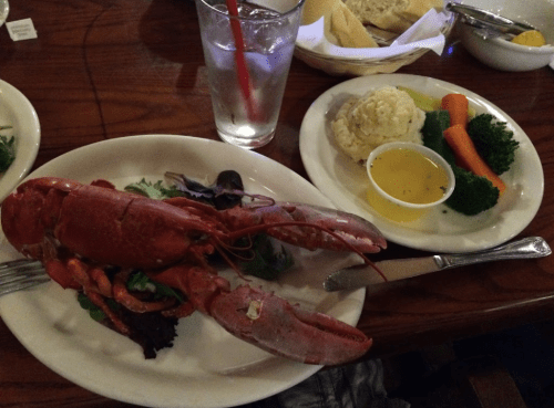 A plate with a whole lobster on greens, a side of mashed potatoes, steamed broccoli, and carrots, with a glass of water.