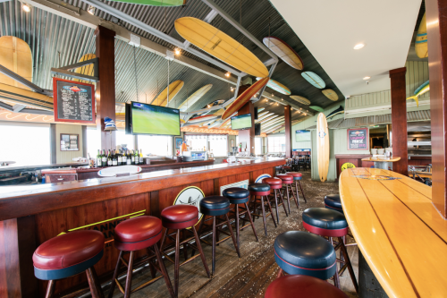 A vibrant bar with surfboards on the ceiling, stools lined up, and TVs displaying sports in a beach-themed setting.