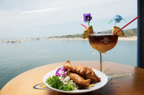 A plate of fried fish with salad beside a colorful cocktail, overlooking a calm harbor with boats in the background.