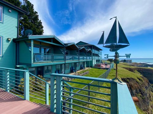 A coastal building with green siding, balconies, and a weather vane shaped like a sailboat overlooking the ocean.