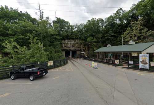 Entrance to a cave with a parking area, surrounded by trees and a building on the right. Cloudy sky above.