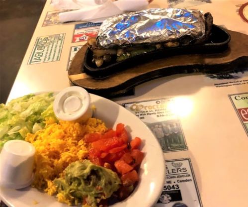 A sizzling fajita wrapped in foil on a wooden platter, with a plate of rice, guacamole, lettuce, and diced tomatoes.