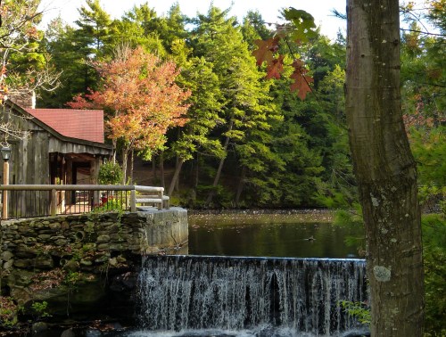 A serene scene of a small waterfall beside a rustic cabin, surrounded by colorful autumn trees and a calm pond.