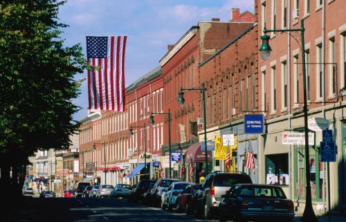 A quaint street lined with brick buildings, featuring an American flag hanging above and parked cars along the curb.