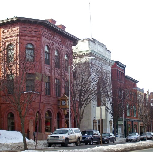 Historic buildings line a snowy street, showcasing red brick architecture and a large archway in the background.