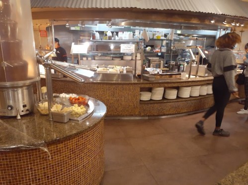 A buffet area with a chocolate fountain, food stations, and a person walking by in a casual setting.