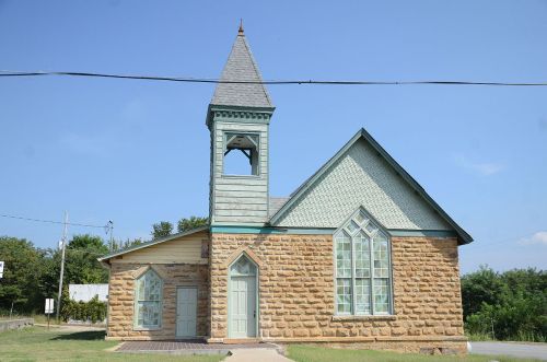 A small stone church with a tall steeple and decorative windows, set against a clear blue sky.