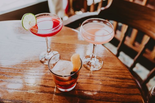 Three colorful cocktails on a wooden table: a red drink with a lime, a pink drink, and a whiskey cocktail with an orange twist.