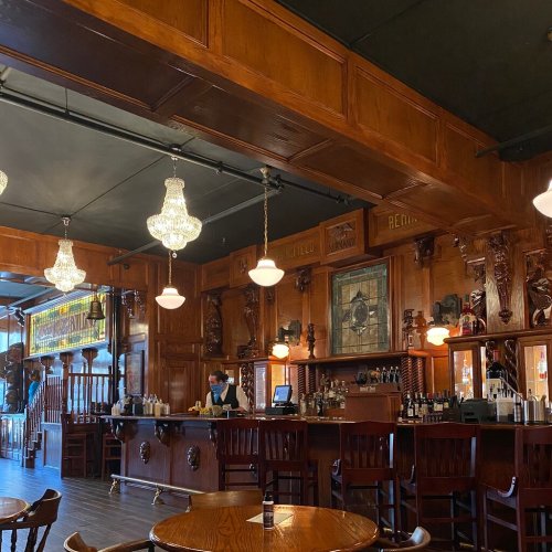 A warm, wooden bar interior with chandeliers, ornate carvings, and a bartender preparing drinks.