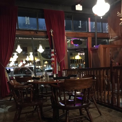 Cozy restaurant interior with wooden tables, red curtains, and hanging lights, featuring an American flag in the background.