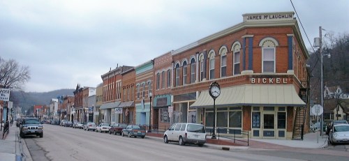 A historic street lined with brick buildings and parked cars under a cloudy sky.