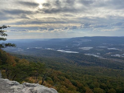 A panoramic view of a valley surrounded by mountains, with cloudy skies and autumn foliage in the foreground.