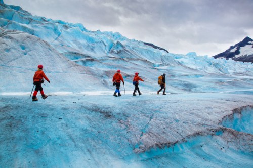 Four climbers in orange jackets trek across a blue glacier under a cloudy sky.