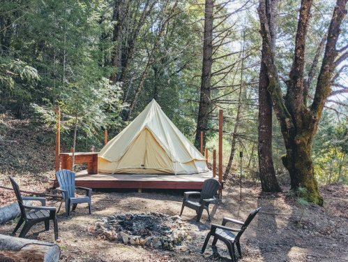 A glamping tent on a wooden platform surrounded by trees, with a fire pit and chairs nearby.