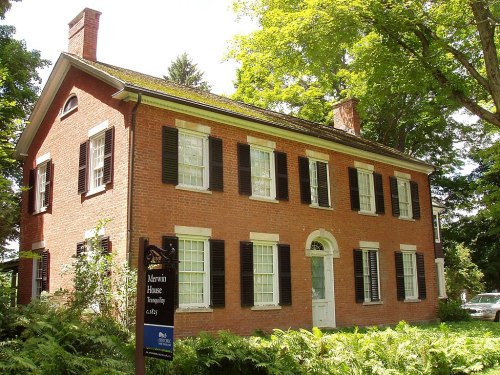 A historic brick house with white trim, surrounded by trees and greenery, featuring a sign for Merwin House.