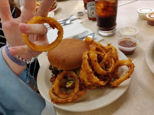A hand holding an onion ring next to a burger and a plate of onion rings, with drinks and condiments in the background.