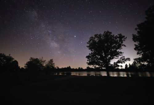 A serene night sky filled with stars and the Milky Way, reflecting over a calm lake surrounded by trees.