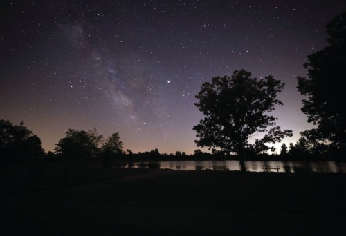 A serene night sky filled with stars and the Milky Way, reflecting over a calm lake surrounded by trees.