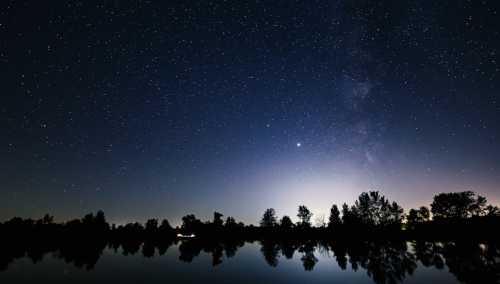 A serene night sky filled with stars reflects on a calm lake, with silhouettes of trees along the horizon.