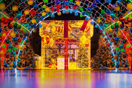 A colorful holiday display featuring bright lights, ornaments, and large gift boxes surrounded by decorated Christmas trees.