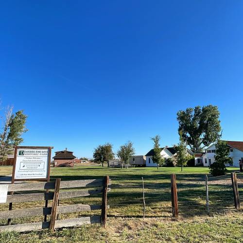 A historic park sign in front of a grassy area with trees and buildings under a clear blue sky.