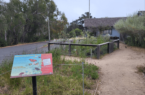 A nature trail sign in front of a building, surrounded by greenery and flowers, on a cloudy day.