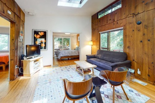 Cozy living area with wooden walls, a sofa, TV, and two chairs, leading to a bedroom with large windows.